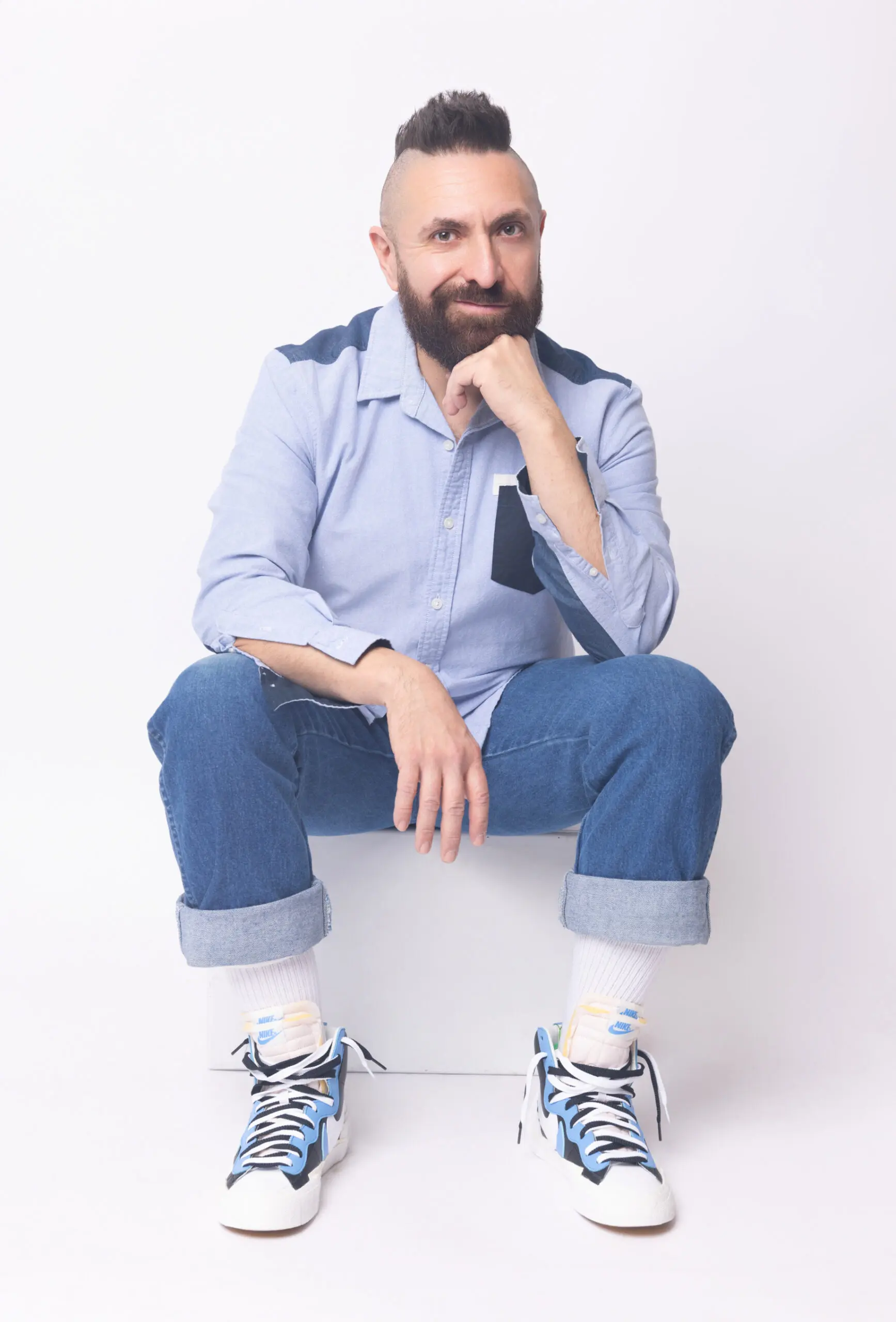 Marcos Vigil, a bearded man with a mohawk, sits casually in a light blue shirt, cuffed jeans, and Nike high-top sneakers against a white background.