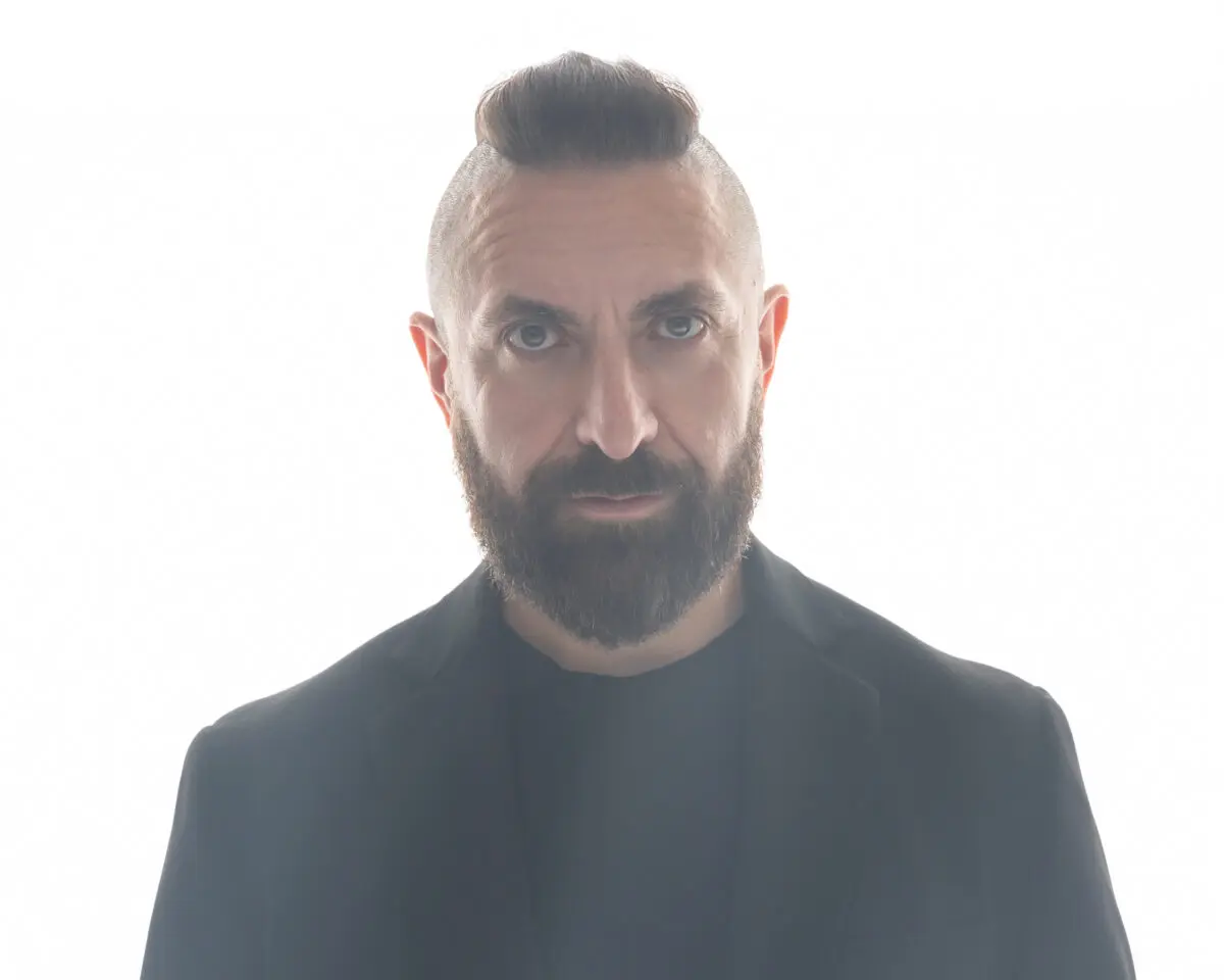 Headshot with Marcos Vigil facing forward with a serious expression, wearing a black blazer, black t-shirt, and his signature mohawk