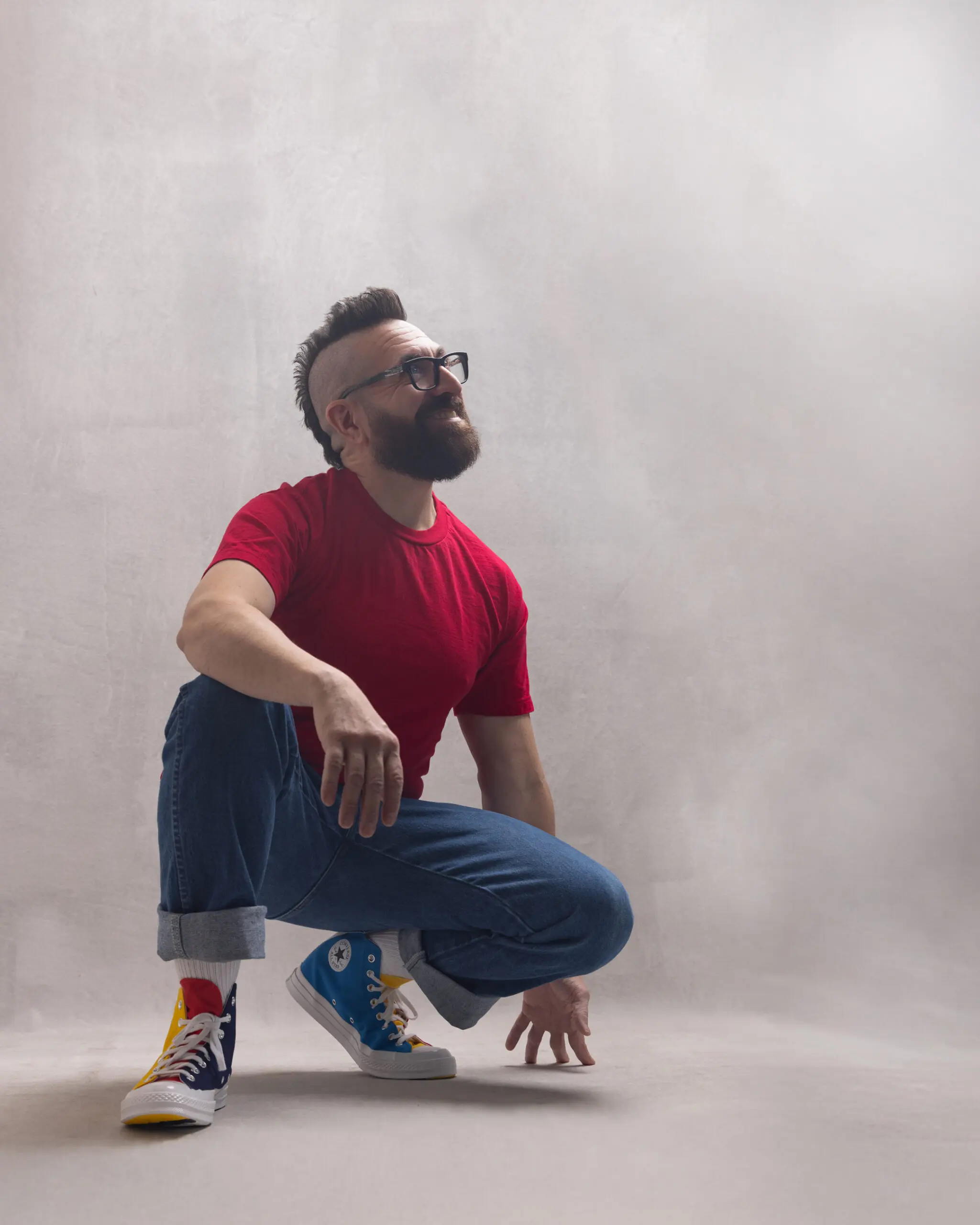 Lyric tenor Marcos Vigil is crouching on the floor with a white background lit from above with studio lighting, looking up towards the light and smiling. He is wearing a bright red t-shirt, cuffed blue jeans, and Converse Chuck Taylor Golf Wang Tri Color Shoes. He has a beard and his signature mohawk.