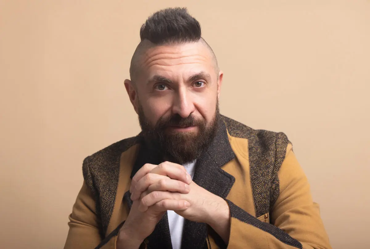 Headshot photo with Marcos Vigil wearing a Junya Wantanabe Blazer with his hands clasped, looking forward at the camera with a serious expression, and he is wearing his signature mohawk.