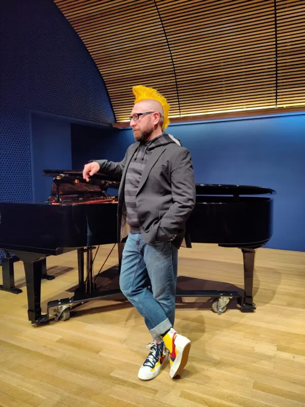 Lyric tenor Marcos Vigil is casually standing in the crook of a black baby grand piano, with his right hand and arm casually resting on the partially open lid of the piano. He is wearing a dark gray blazer, a Junya Watanabe gray hoodie with dark gray horizontal stripes and reflector details on the hood, blue jeans that are cuffed, and a pair of Sacai Blazer Mid sneakers in the red and yellow colorway. He is wearing a beard, glasses, and his signature mohwak dyed yellow and spiked up.
