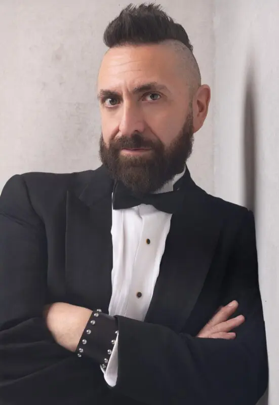Marcos Vigil is standing with his arms crossed while leaning against a wall on his left side. He has a beard and a mohawk, and is wearing a formal tuxedo with a black tie, black buttons with gold trim,, and on his left wrist he is wearing a black leather cuff with round silver metal studs on the edges of the cuff.