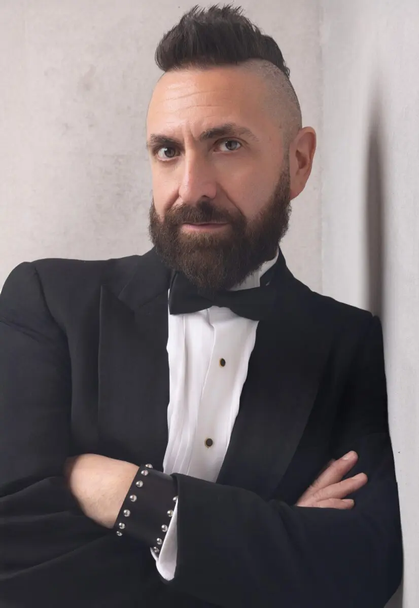 Headshot photo of Marcos Vigil wearing a black tuxedo jacket and black bow tie, arms crossed leaning against a white background, showing a black leather cuff with silver studs on his left wrist, looking forward with a serious expression, wearing his signature mohawk.