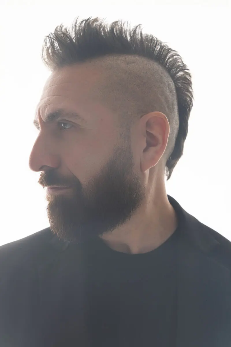 Headshot photo of Marcos Vigil at profile, wearing a black blazer, black t-shirt, and a serious expression, wearing his signature mohawk spiked up. 