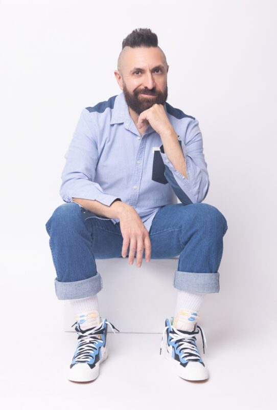 Marcos Vigil, a bearded man with a mohawk, sits casually in a light blue shirt, cuffed jeans, and Nike high-top sneakers against a white background.