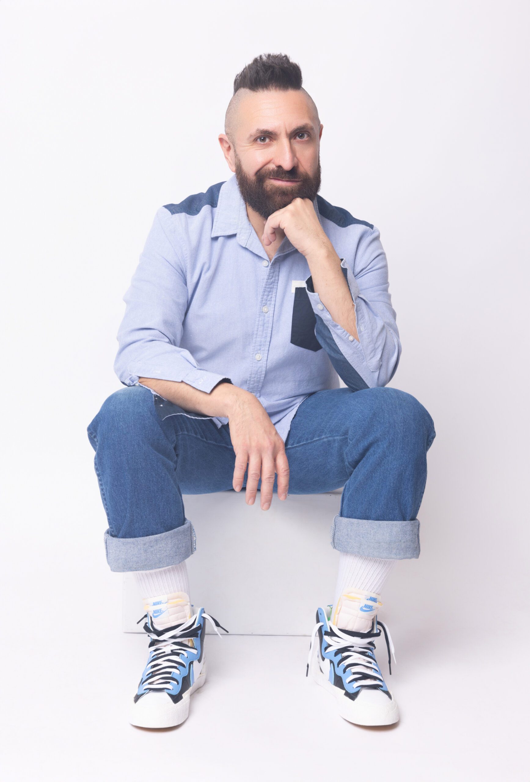 Marcos Vigil, a bearded man with a mohawk, sits casually in a light blue shirt, cuffed jeans, and Nike high-top sneakers against a white background.