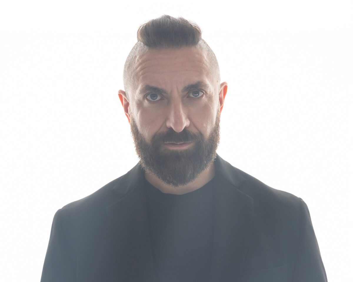 Headshot with Marcos Vigil facing forward with a serious expression, wearing a black blazer, black t-shirt, and his signature mohawk