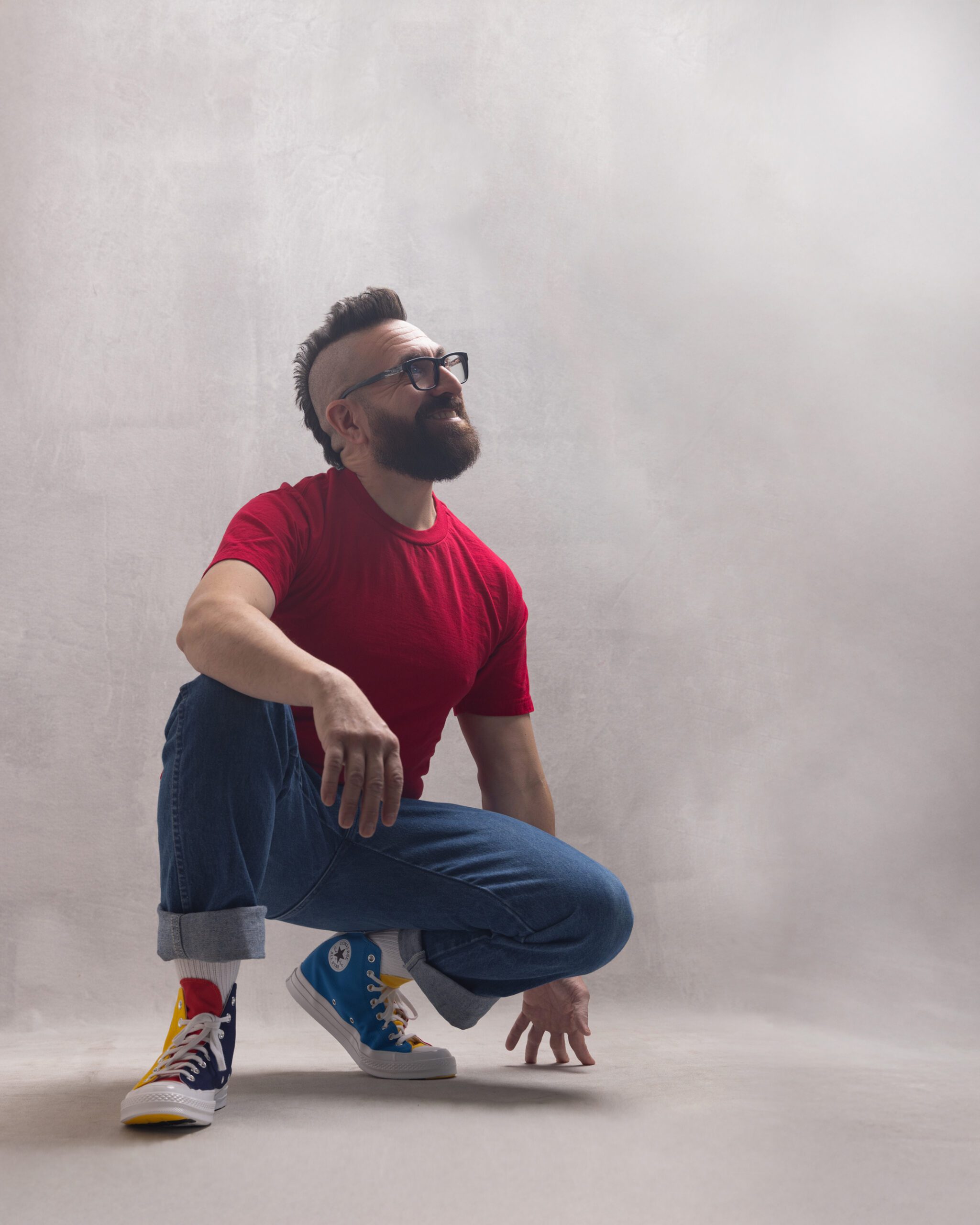 Lyric tenor Marcos Vigil is crouching on the floor with a white background lit from above with studio lighting, looking up towards the light and smiling. He is wearing a bright red t-shirt, cuffed blue jeans, and Converse Chuck Taylor Golf Wang Tri Color Shoes. He has a beard and his signature mohawk.