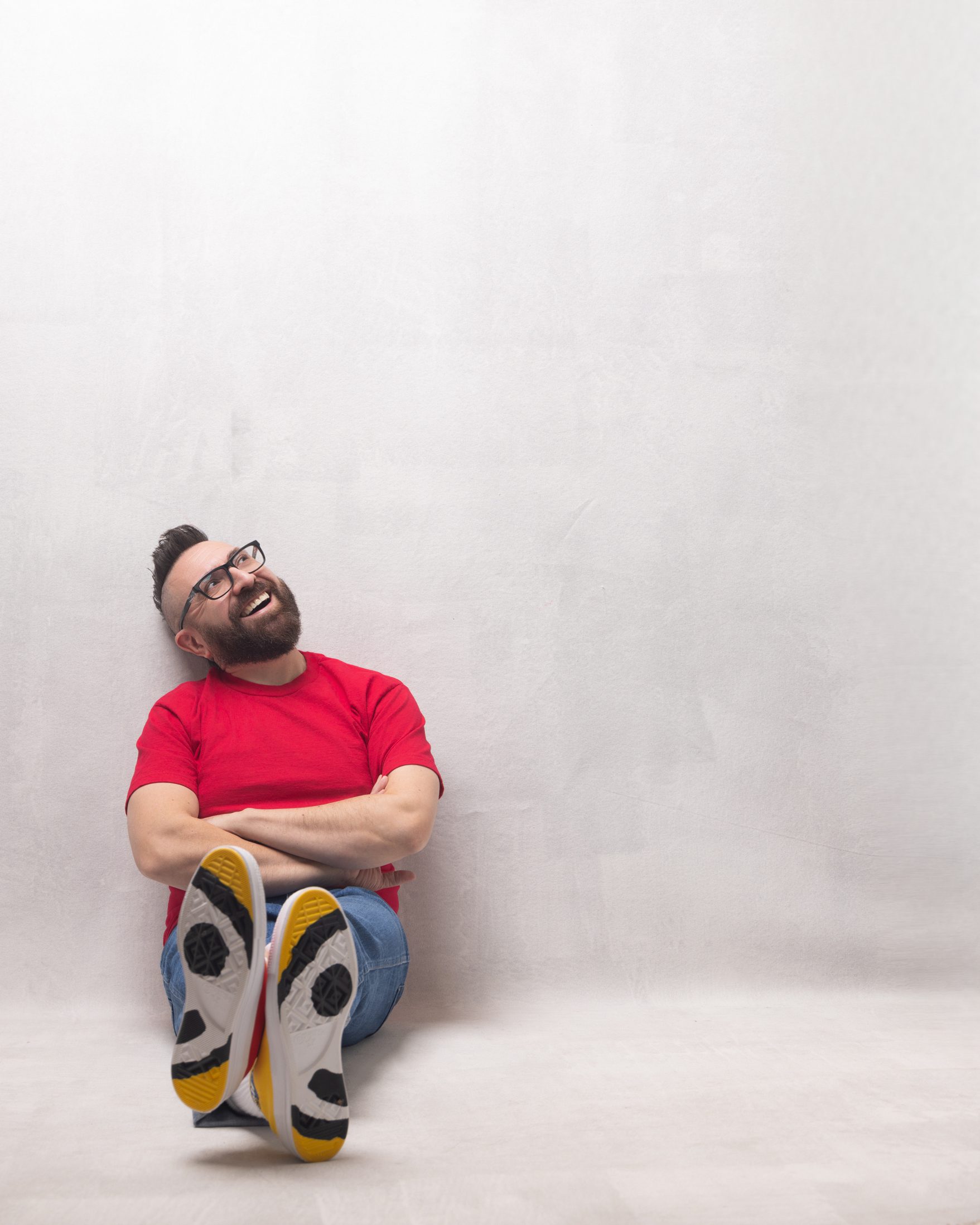 Lyric Tenor Marcos Vigil is sitting on the white floor of a studio with a white background his arms are crossed and his legs are extended to the front so that the viewer can see the soles of his shoes, and his feet are crossed. HE is wearing a bright red t-shirt, cuffed blue jeans and Converse Chuck Taylor Golf Wang Tripanel shoes. He is smiling while looking up with his head slightly tilted to the right. He is wearing glasses, has a beard, and his signature mohawk.