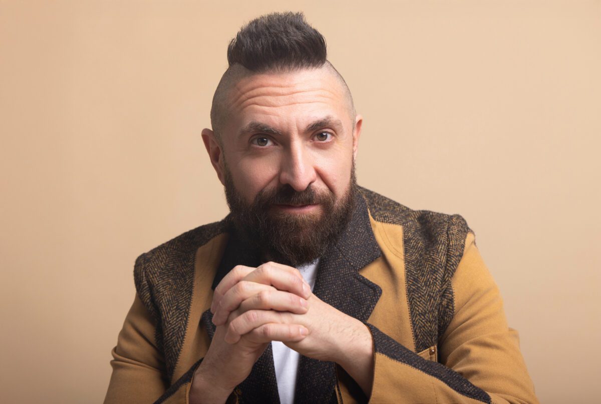 Headshot photo with Marcos Vigil wearing a Junya Wantanabe Blazer with his hands clasped, looking forward at the camera with a serious expression, and he is wearing his signature mohawk.