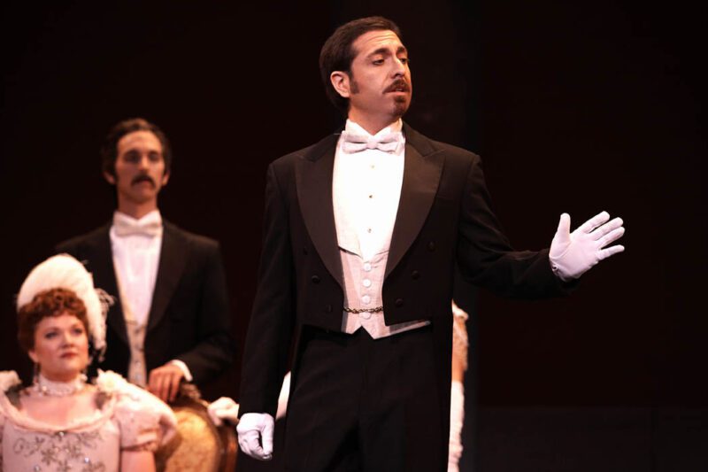Marcos Vigil, tenor, is wearing a formal tuxedo with a white waist coat and a white bow tie, white gloves, and a pocket watch. he has short brown hair and a moustache. He is standing in front of party guests while looking to the left and gesturing with his left hand.