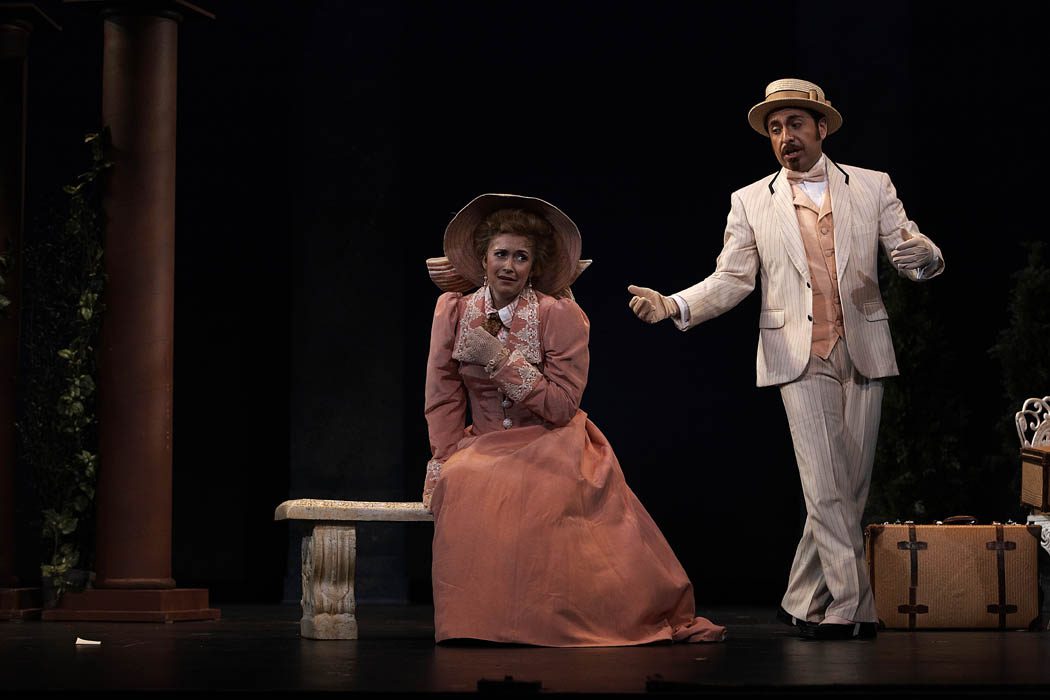 Marcos Vigil, Tenor, is wearing a light colored pin stripe suite, with a peach colored waist coat and matching bow tie, light colored gloves, and a boater hat.