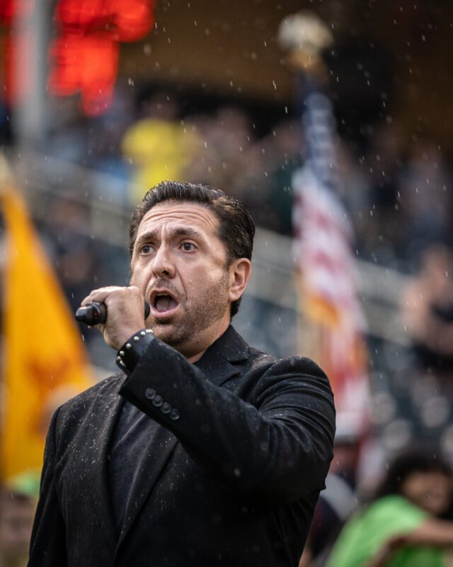 Marcos Vigil, tenor, sings the National Anthem in the rain holding a microphone with the New Mexican and American flags waving in the background.