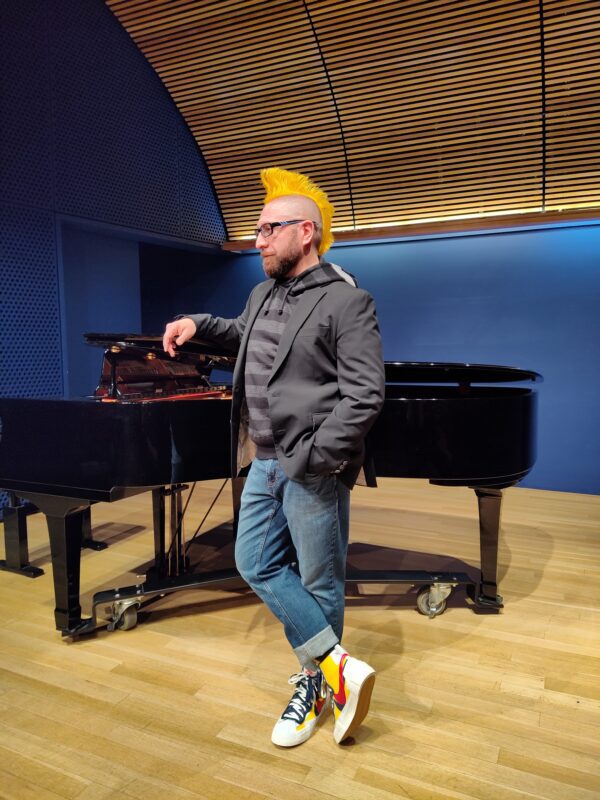Lyric tenor Marcos Vigil is casually standing in the crook of a black baby grand piano, with his right hand and arm casually resting on the partially open lid of the piano. He is wearing a dark gray blazer, a Junya Watanabe gray hoodie with dark gray horizontal stripes and reflector details on the hood, blue jeans that are cuffed, and a pair of Sacai Blazer Mid sneakers in the red and yellow colorway. He is wearing a beard, glasses, and his signature mohwak dyed yellow and spiked up.