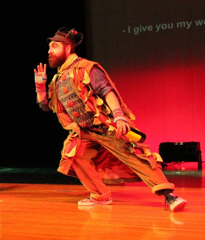 Marcos Vigil, Tenor, wearing a very colorful costume with colorful knit finger-less gloves, holding a bottle of wine in his left hand while leaning deep towards the right, on stage with with red stage lighting in the background.