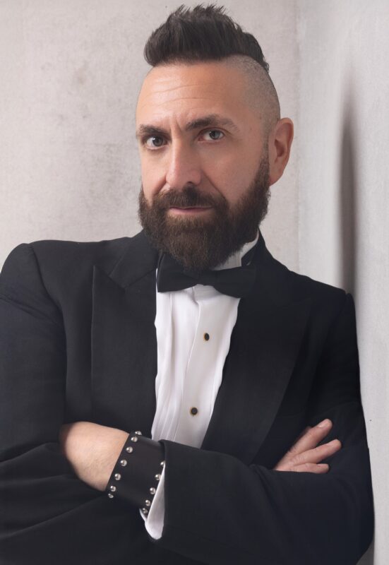 Marcos Vigil is standing with his arms crossed while leaning against a wall on his left side. He has a beard and a mohawk, and is wearing a formal tuxedo with a black tie, black buttons with gold trim,, and on his left wrist he is wearing a black leather cuff with round silver metal studs on the edges of the cuff.