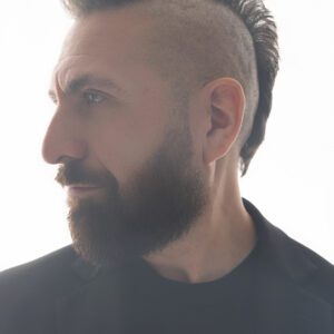 Marcos Vigil, Tenor, is standing with his shoulders facing forward while his face is turned to his right, looking off into the distance so that his mohawk is viewed in profile. He is wearing a black blazer with a black t-shirt.