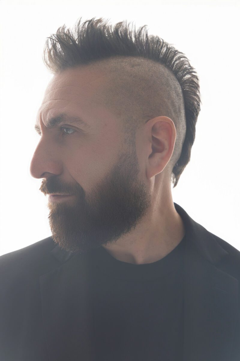 Headshot photo of Marcos Vigil at profile, wearing a black blazer, black t-shirt, and a serious expression, wearing his signature mohawk spiked up. 