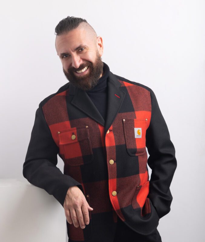 Marcos Vigil, tenor, standing with one hand in his pocket and the other arm placed on a white platform. He is wearing a Junya Watanabe red and black blazer with a Carhartt logo on the upper left pocket, a black turtle neck, and black jeans.